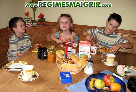 Trois jeunes enfants sont en train de prendre le petit-déj' dans la bonne humeur autour d'une table en bois