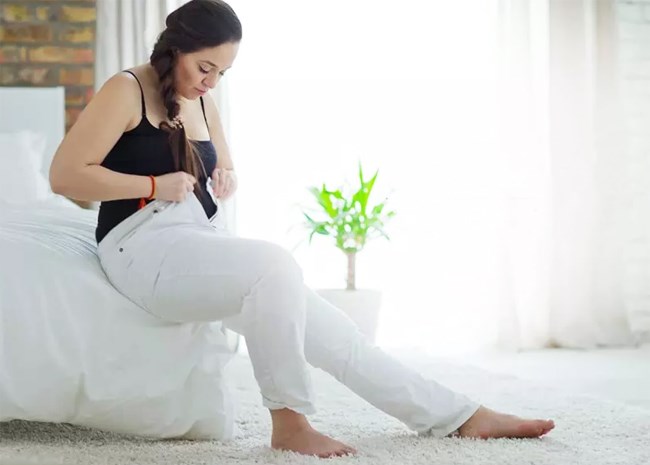 Une femme en surcharge pondrale tente de boutonner son pantalon en s'asseyant sur un lit