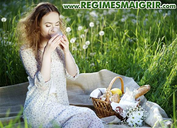 Une femme boit un verre de lait assise sur une nappe sur laquelle se trouve un panier pique-nique